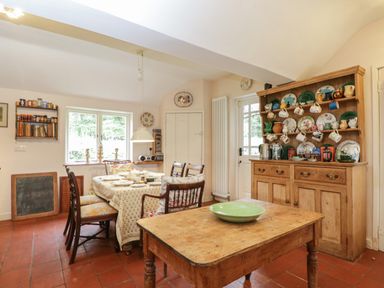 A dining room with a wooden table set for meals chairs a wooden cupboard with dishes and mugs and a window at Chalkpit Cottage in Blewbury