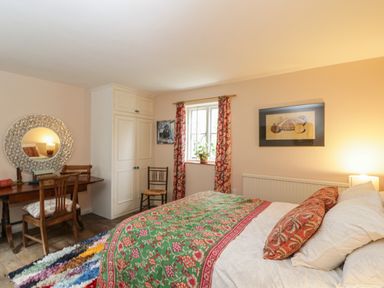 A bedroom with a bed covered by a patterned green and red blanket a wooden desk with a round mirror and chairs a window with floral curtains at Chalkpit Cottage in Blewbury