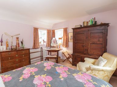 A bedroom with a wooden dresser, vanity table with mirror, armchair, wardrobe, and bed with floral bedspread at Chalkpit Cottage in Blewbury