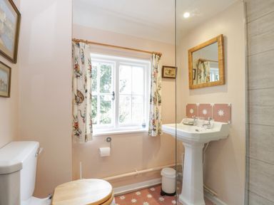 A bathroom with a toilet with wooden seat a pedestal sink a mirror window with patterned curtains and a tiled floor at Chalkpit Cottage in Blewbury