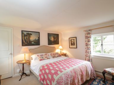 A bedroom with a double bed covered with a pink patterned blanket two side tables with lamps and paintings on walls at Chalkpit Cottage in Blewbury