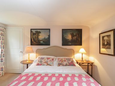 A bedroom with a bed with pillows and lamps on side tables and paintings on the walls at Chalkpit Cottage in Blewbury