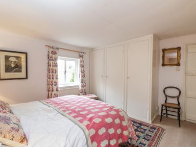 A bedroom with a bed covered with pink and white blankets a window with floral curtains built in white wardrobes a black chair with a cane seat a mirror and a framed picture on the walls at Chalkpit Cottage in Blewbury