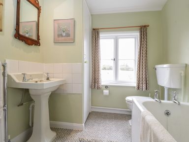 A bathroom with a pedestal sink, toilet, bathtub, window with curtains, and a mirror at Chalkpit Cottage in Blewbury