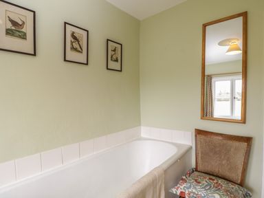 A bathroom with a white bathtub a beige towel hanging on the tub bird pictures on the wall a wooden chair with a patterned cushion and a wall mirror at Chalkpit Cottage in Blewbury