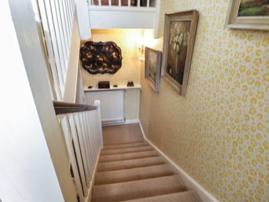 A staircase with beige carpet leading down with yellow patterned wallpaper and framed paintings on the wall at Chalkpit Cottage in Blewbury