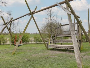 A wooden playground structure with a swing and steps on grass with trees and fields in the background at Chalkpit Cottage in Blewbury