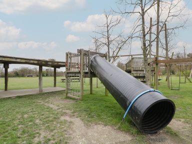 An outdoor playground with a large black tube slide wooden climbing structure and swings in a grassy area