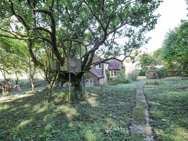 A garden with a treehouse and a brick house in the background at Chalkpit Cottage in Blewbury