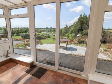 A sunroom with tiled floor and glass doors leading to a garden with patio table and landscaped greenery at Pen Bonc  Llanddaniel Near Gaerwen