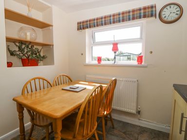 A dining area with a wooden table and four chairs near a window with red decorative items and a wall clock at Pen Bonc in Llanddaniel Near Gaerwen