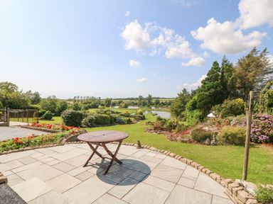 A patio with a wooden table overlooking a garden with shrubs trees and a pond at Pen Bonc in Llanddaniel Near Gaerwen
