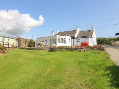 A single story white house with a glass sunroom next to a stone wall garden with flowers and propane tanks at Pen Bonc in Llanddaniel Near Gaerwen