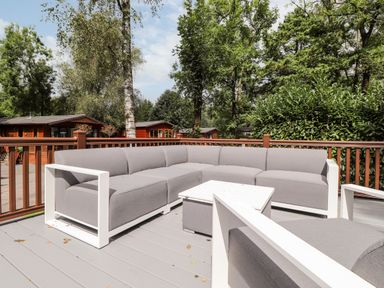 An outdoor deck with grey cushioned sectional sofa white framed armchairs and a white coffee table surrounded by trees at Unique Luxury Lodge 1 Langdale White Cross Bay near Windermere