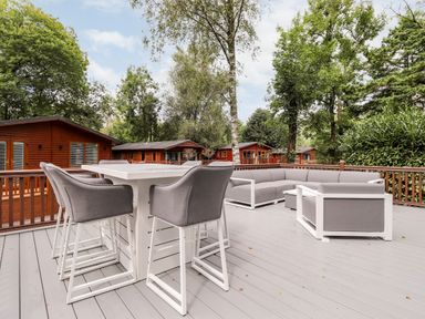 An outdoor deck with a high dining table with chairs and a sectional sofa with trees and wooden cabins in the background at Unique Luxury Lodge 1 Langdale White Cross Bay near Windermere