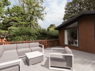 An outdoor deck area with a gray sectional sofa armchair and a small table next to a brown wooden building at Unique Luxury Lodge 1 Langdale White Cross Bay near Windermere