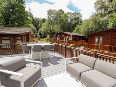 A deck with outdoor seating and dining area overlooking wooden lodges surrounded by trees at Unique Luxury Lodge 1 Langdale White Cross Bay near Windermere