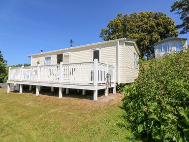 An exterior view of a mobile home with a white deck and railing on a grassy slope with shrubs and trees at Horizon in Combe Martin