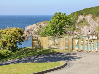 A fenced outdoor pool area with white chairs near a grassy slope overlooking the sea and rocky cliffs at Horizon in Combe Martin