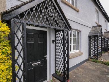 A black door with a lattice porch and window on the side of a light grey building at Hobbs Cottage in Sticklepath