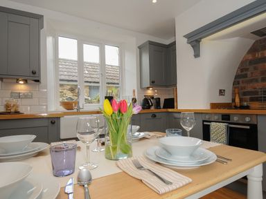 A kitchen with grey cabinets a wooden countertop a dining table set with white dishes glasses and a vase with tulips at Hobbs Cottage in Sticklepath
