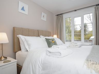 A bedroom with a bed with white linens lamps on either side and a window with curtains at Hobbs Cottage in Sticklepath