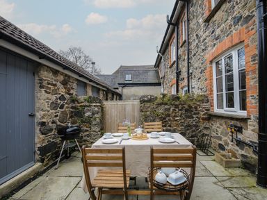 An outdoor stone patio with wooden dining table and chairs set with dishes and cups next to a stone building at Hobbs Cottage in Sticklepath