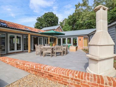 A patio area with wooden dining table and chairs under a green umbrella next to a chimney at Church Farm Barn Dunwich near Westleton