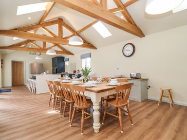 A dining area with a wooden table and chairs set with dishes and glasses next to a kitchen island with stools under exposed wooden ceiling beams at Church Farm Barn Dunwich near Westleton