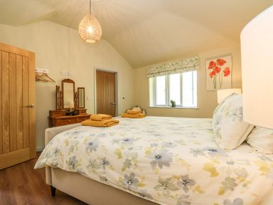 A bedroom with a bed covered in floral bedding towels on the bed a wooden dresser with a mirror a window with a patterned curtain and a floral painting on the wall at Church Farm Barn Dunwich near Westleton