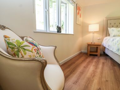 A bedroom with a white armchair with printed cushions beside a window and a wooden side table with a lamp at Church Farm Barn Dunwich near Westleton