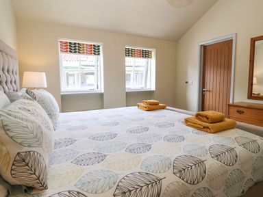 A bedroom with a bed covered in a leaf-patterned duvet two windows with patterned blinds a bedside lamp a wooden door and a wooden dresser with a mirror at Church Farm Barn in Dunwich near Westleton
