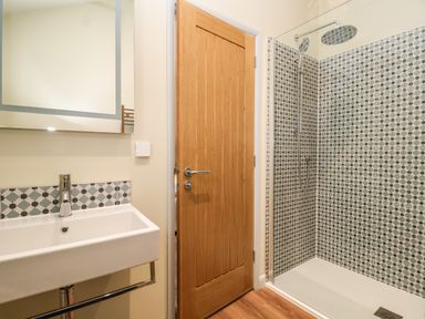 A bathroom with a sink and mirror on the left a wooden door in the center and a shower with patterned tiles on the right at Church Farm Barn in Dunwich near Westleton