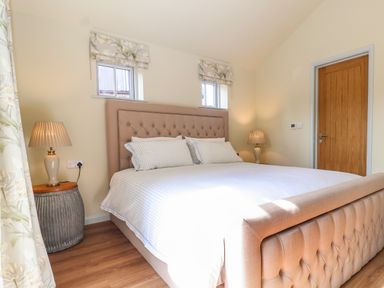 A bedroom with a beige upholstered bed two bedside tables with lamps and wooden door at Church Farm Barn in Dunwich near Westleton