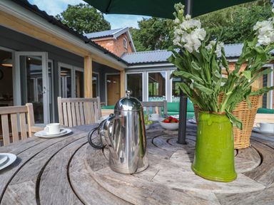 An outdoor wooden table with a metal coffee pot a green vase with white flowers cups and bowls of fruit at Church Farm Barn in Dunwich near Westleton