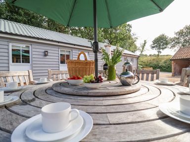 An outdoor wooden table with white cups plates bowls of fruit a basket a teapot and flowers under a green umbrella at Church Farm Barn Dunwich near Westleton