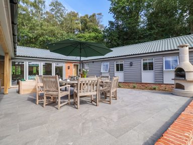 An outdoor patio area with wooden dining table and chairs under a green umbrella and a stone barbecue at Church Farm Barn in Dunwich near Westleton