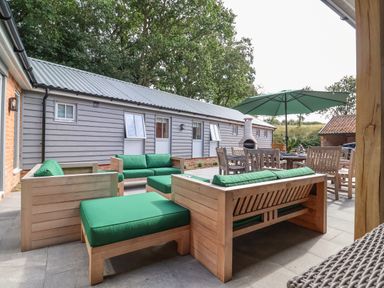 An outdoor patio with wooden seating and green cushions a dining table with chairs and an umbrella at Church Farm Barn in Dunwich near Westleton