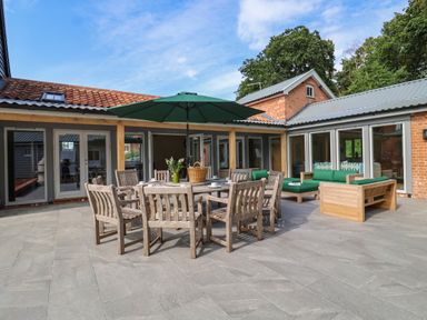 An outdoor patio area with a wooden dining table and chairs under a green umbrella and wooden sofas with green cushions at Church Farm Barn in Dunwich near Westleton