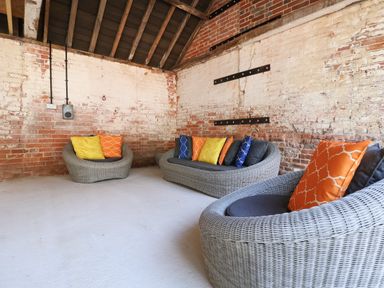 A room with exposed brick walls and ceiling beams containing three gray wicker sofas with colorful cushions at Church Farm Barn Dunwich near Westleton