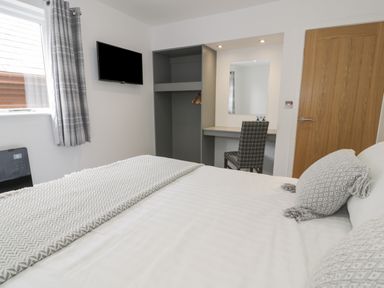 A bedroom with a bed white bedding throw pillows a wall mounted tv a window with curtains a wooden door a gray open wardrobe and a desk with a chair at Classic Lodge 16 Dyserth