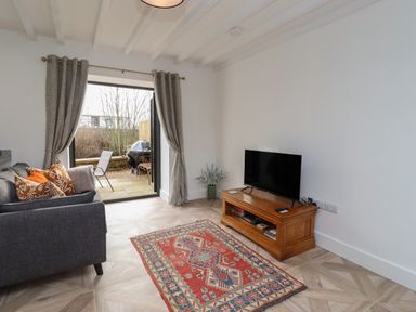 A living room with a gray sofa orange and patterned cushions a wooden TV stand with a television a patterned rug and open doors to a patio at Rowan Barn in Bellingham