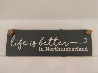 A black wooden sign with white text that says life is better in Northumberland hanging on a white wall at Rowan Barn in Bellingham