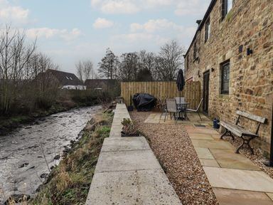 A stone patio with a bench table and chairs next to a river by a stone building at Rowan Barn in Bellingham
