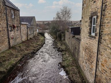A narrow river flowing between stone buildings with a small bridge in the background at Rowan Barn in Bellingham