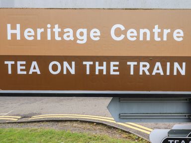 A brown and beige sign with the text Heritage Centre tea on the train by a road with double yellow lines at Rowan Barn in Bellingham