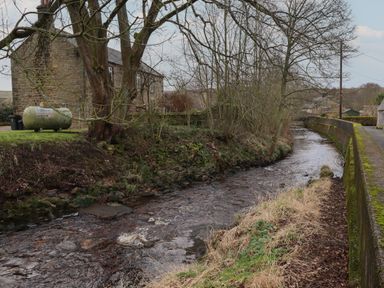 A flowing stream beside a stone house and trees with a mossy wall and pathway at Rowan Barn in Bellingham