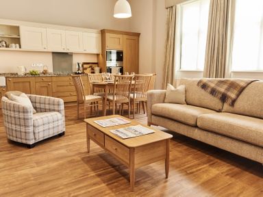 A living room with a sofa a chair a wooden coffee table with newspapers and an open kitchen with a dining table and chairs at The Stalls in Humshaugh