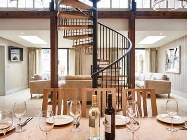 A dining table set with glasses plates and wine bottles in a room with spiral staircase and sofas at The Clocktower in Humshaugh