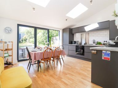 A kitchen and dining area with wooden chairs around a table covered with a red tablecloth near glass doors opening to a garden at Long View in Southwold